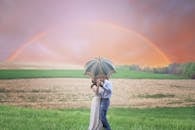 A couple shares a kiss under an umbrella in a scenic rural landscape with a rainbow.